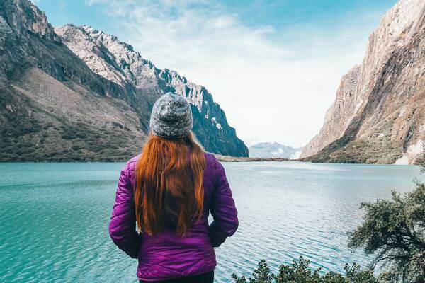Eine Frau in einer violetten Jacke und einer grauen Mütze steht an einem türkisblauen See mit Bergen im Hintergrund.