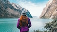 A woman in a purple jacket and gray beanie standing by a turquoise lake with mountains in the background.