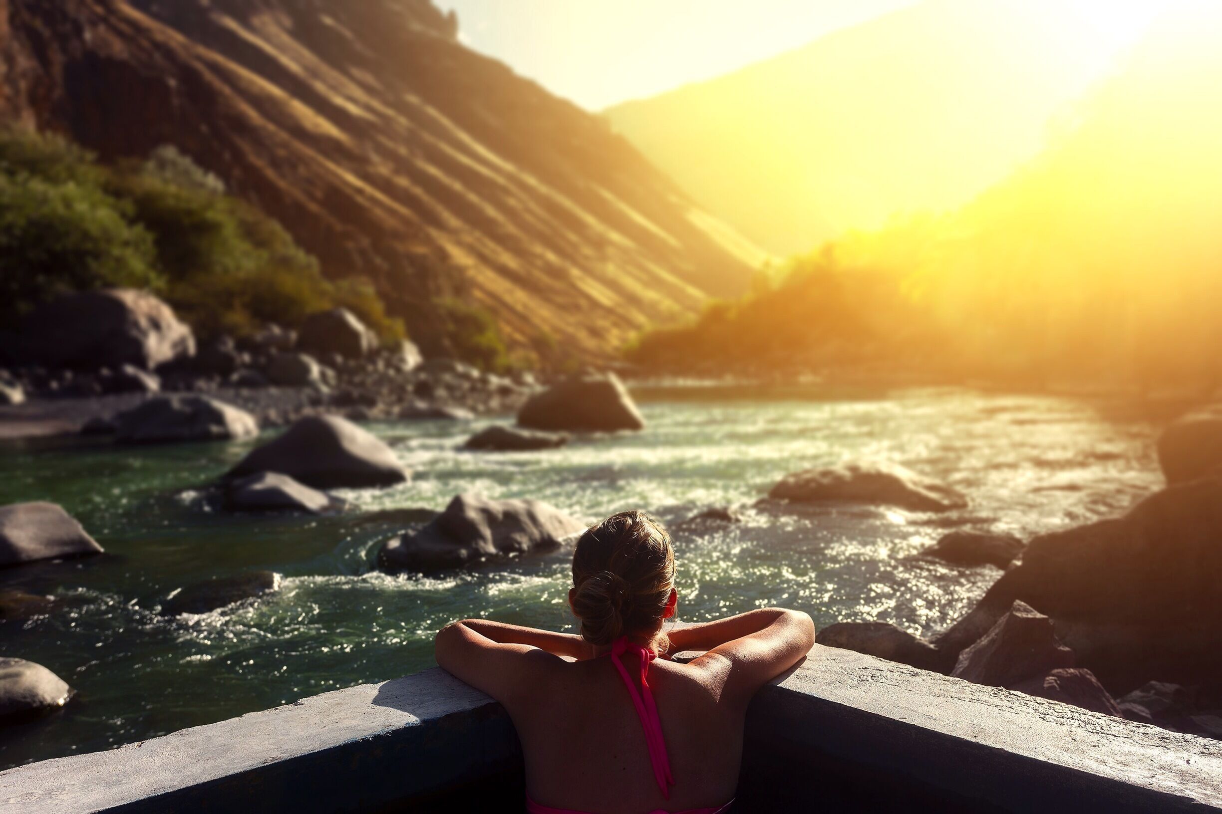 A person in a pink swimsuit relaxing by a river with rocks and hills in the background.