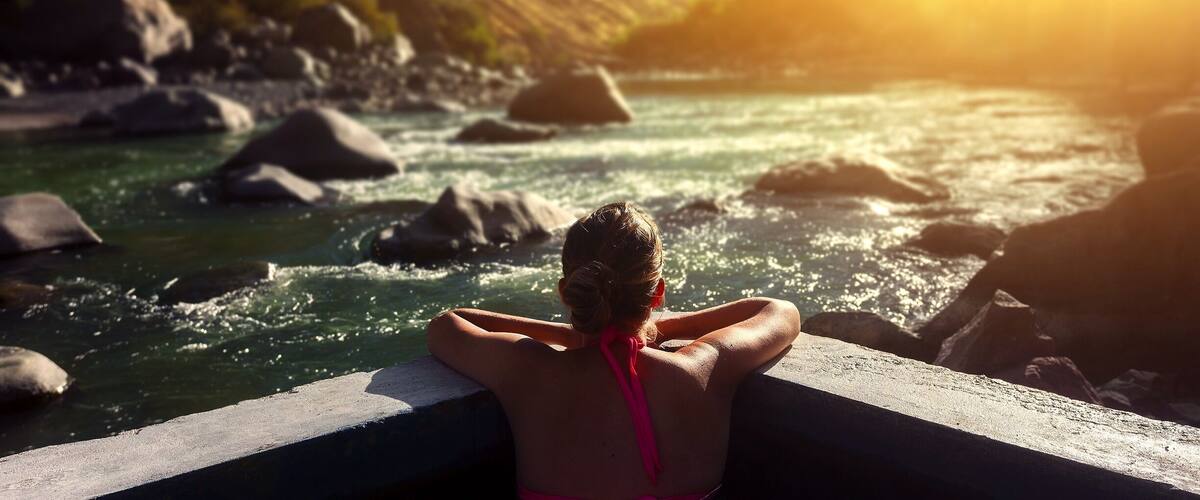 A person in a pink swimsuit relaxing by a river with rocks and hills in the background.