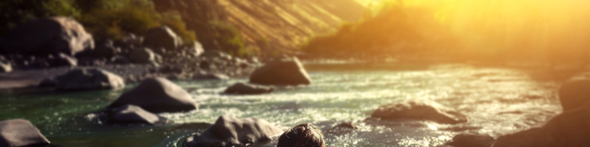 A person in a pink swimsuit relaxing by a river with rocks and hills in the background.