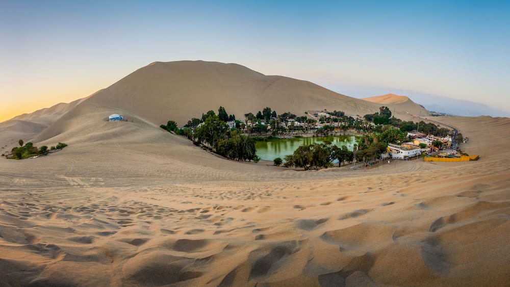 A desert landscape with sand dunes, a small oasis with a pond, and a settlement with buildings.