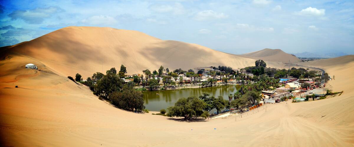 A desert landscape with sand dunes, a small lake, and a village with buildings and trees.