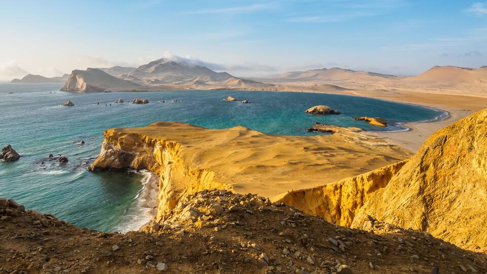 A coastal landscape with a prominent cliff, turquoise sea, and distant mountains.
