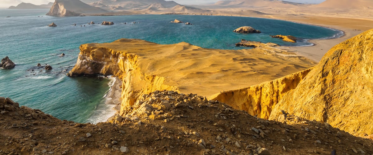 A coastal landscape with a prominent cliff, turquoise sea, and distant mountains.
