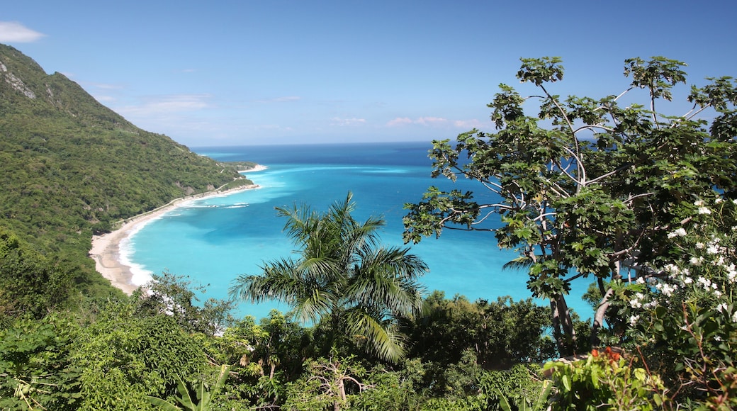 Paisagem costeira com mar azul límpido, praia arenosa e colinas verdejantes.
