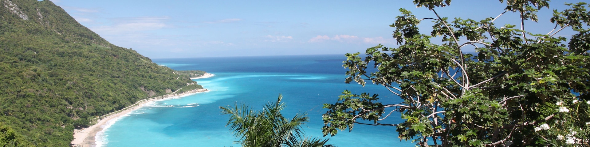A coastal landscape with a clear blue sea, a sandy beach, and lush green hills.