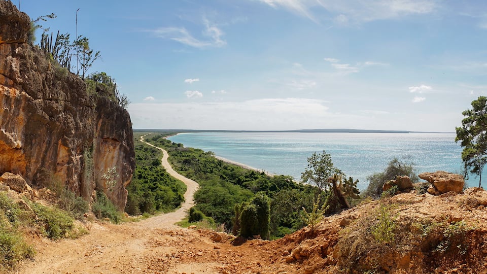 A coastal landscape with a dirt path leading to a beach, surrounded by rocky cliffs and sparse vegetation.