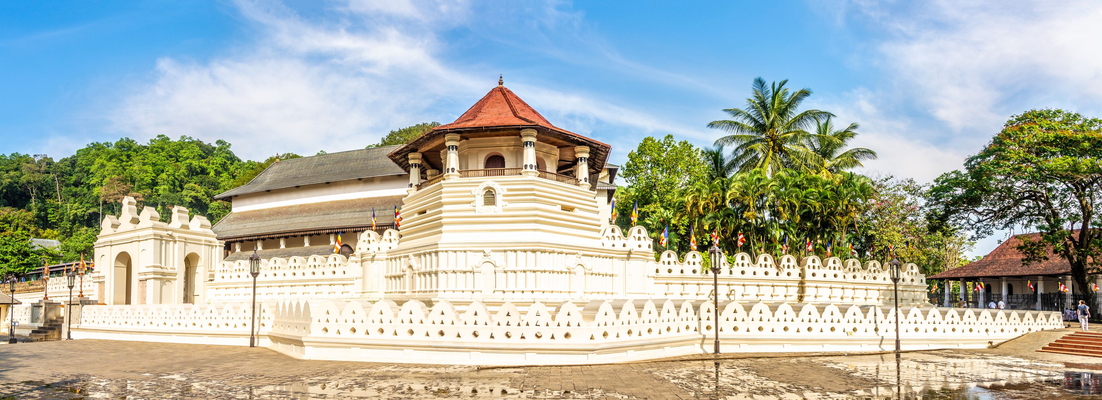 A large white temple with a red roof, surrounded by lush greenery and palm trees.