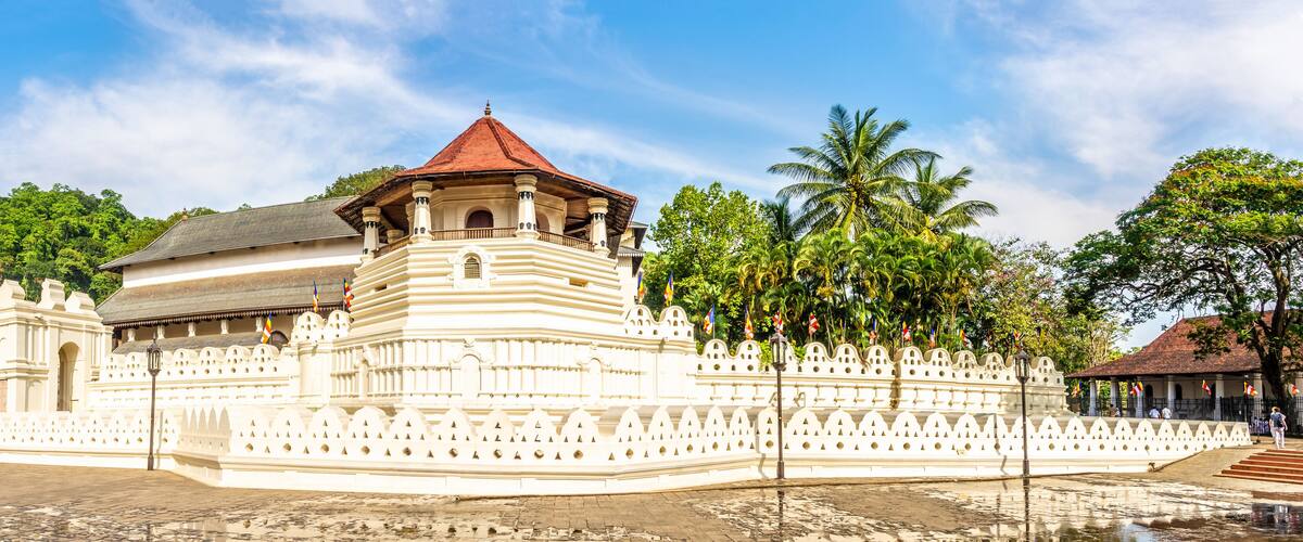 A large white temple with a red roof, surrounded by lush greenery and palm trees.