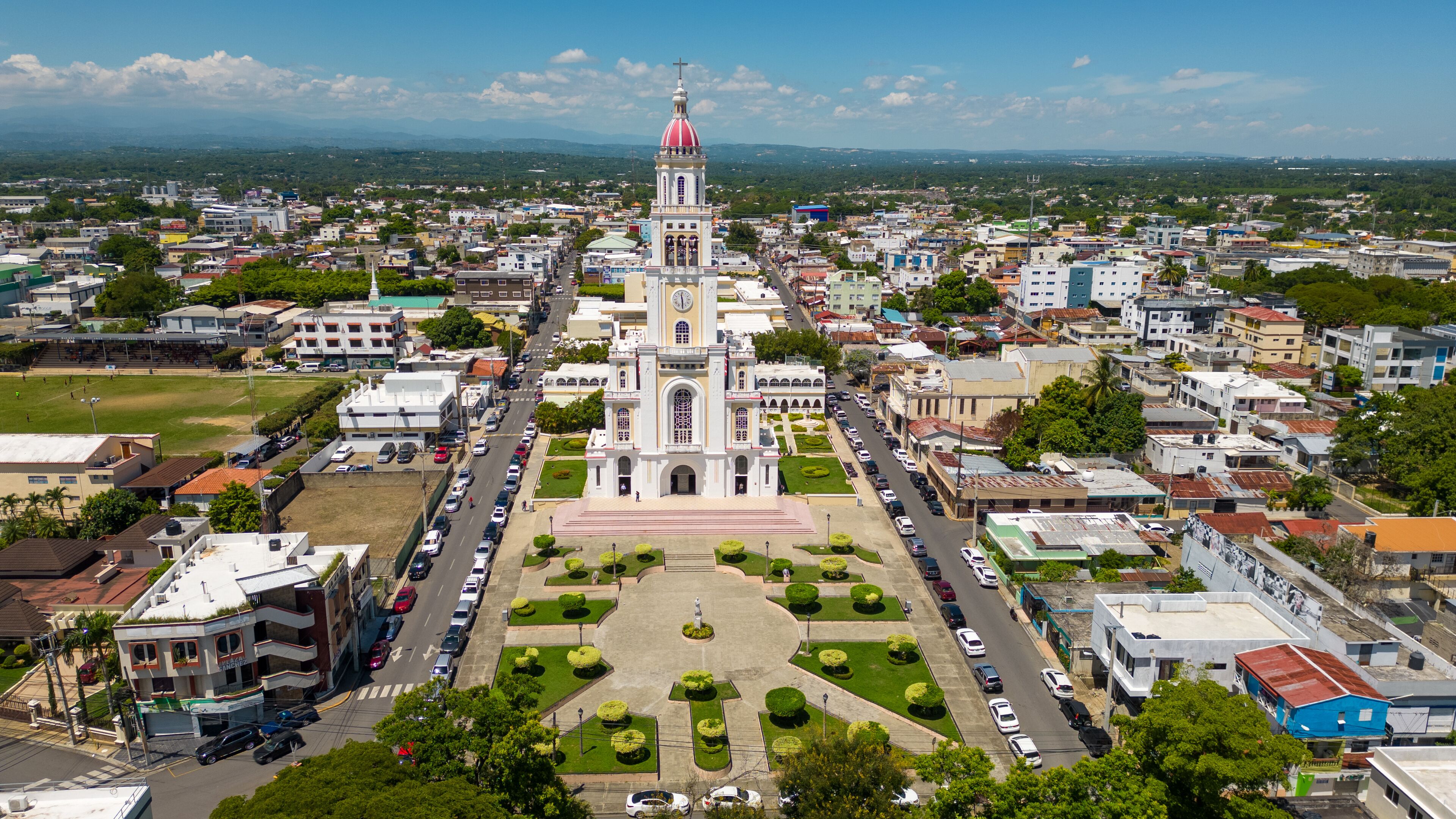 An aerial view of a city with a prominent church featuring a red dome, surrounded by a mix of residential and commercial buildings, and a green park in the center.