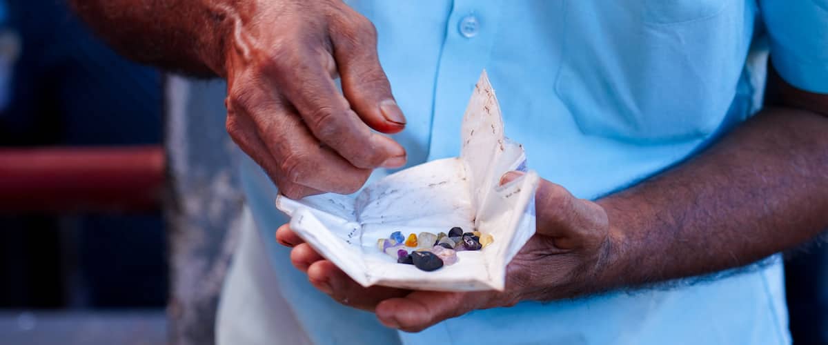 An individual at the Nivithigala gem market in Sri Lanka presents a collection of rough gemstones wrapped in paper for evaluation. He is negotiating with another person to secure a better price.