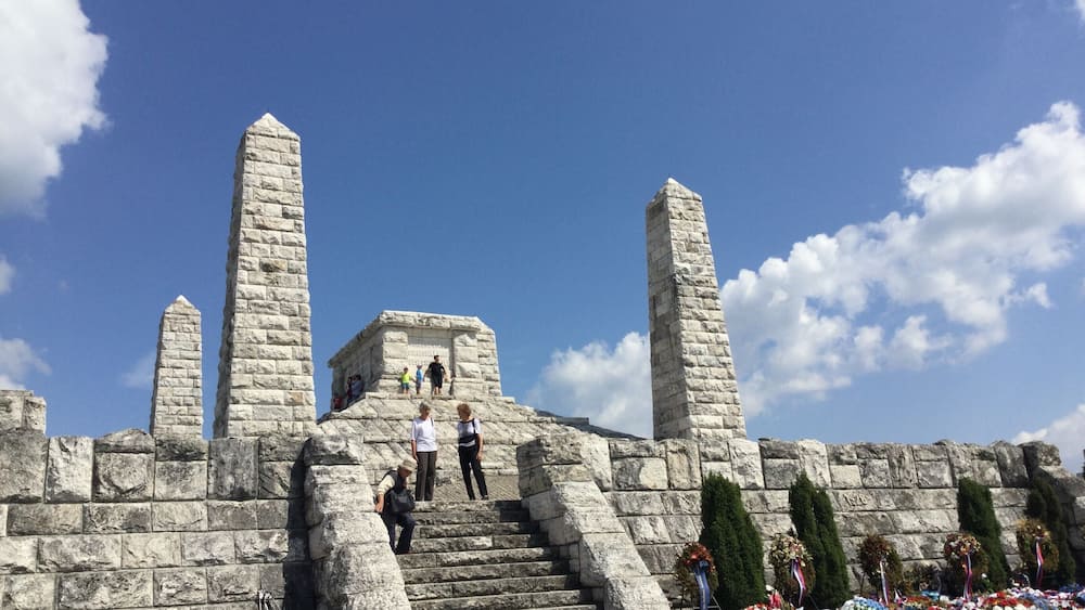 Un monument de pierre orné d’inscriptions, un escalier menant à celui-ci et un espace commémoratif décoré de guirlandes.