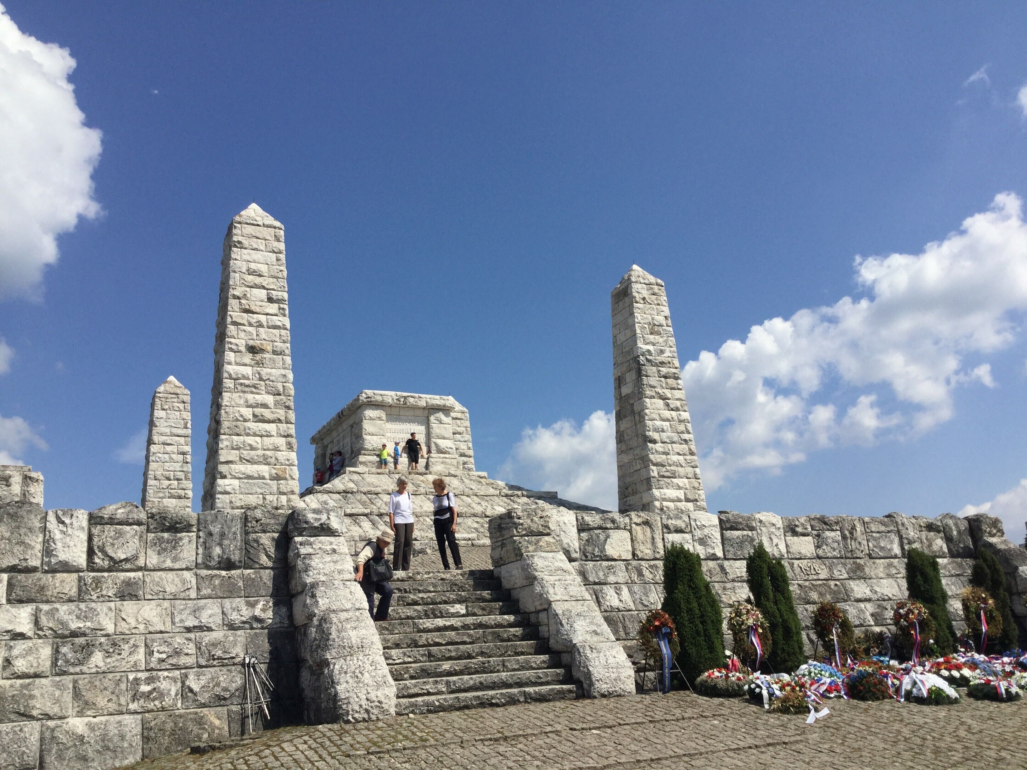 A stone monument with inscriptions, a staircase leading up to it, and a wreath-laden memorial area.