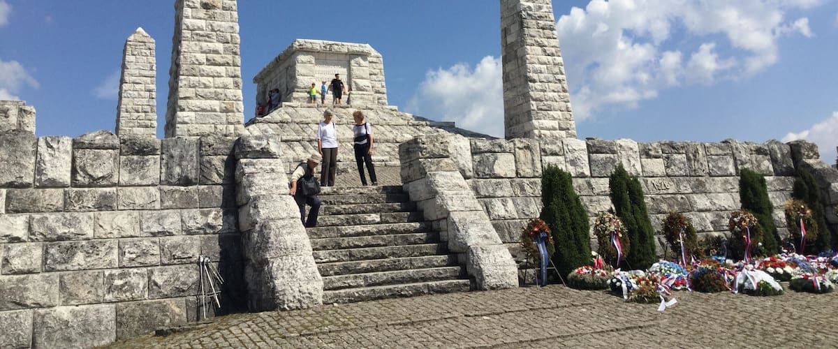 A stone monument with inscriptions, a staircase leading up to it, and a wreath-laden memorial area.