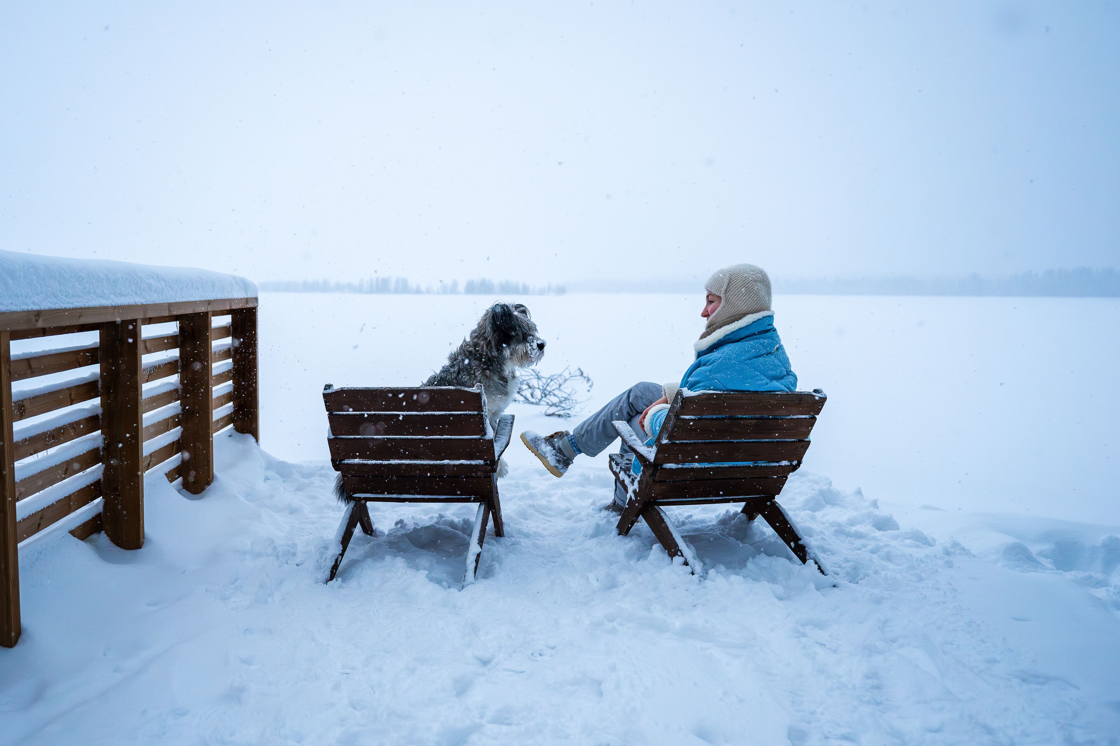 woman and her dog sit on wooden chairs near snowy frozen lake