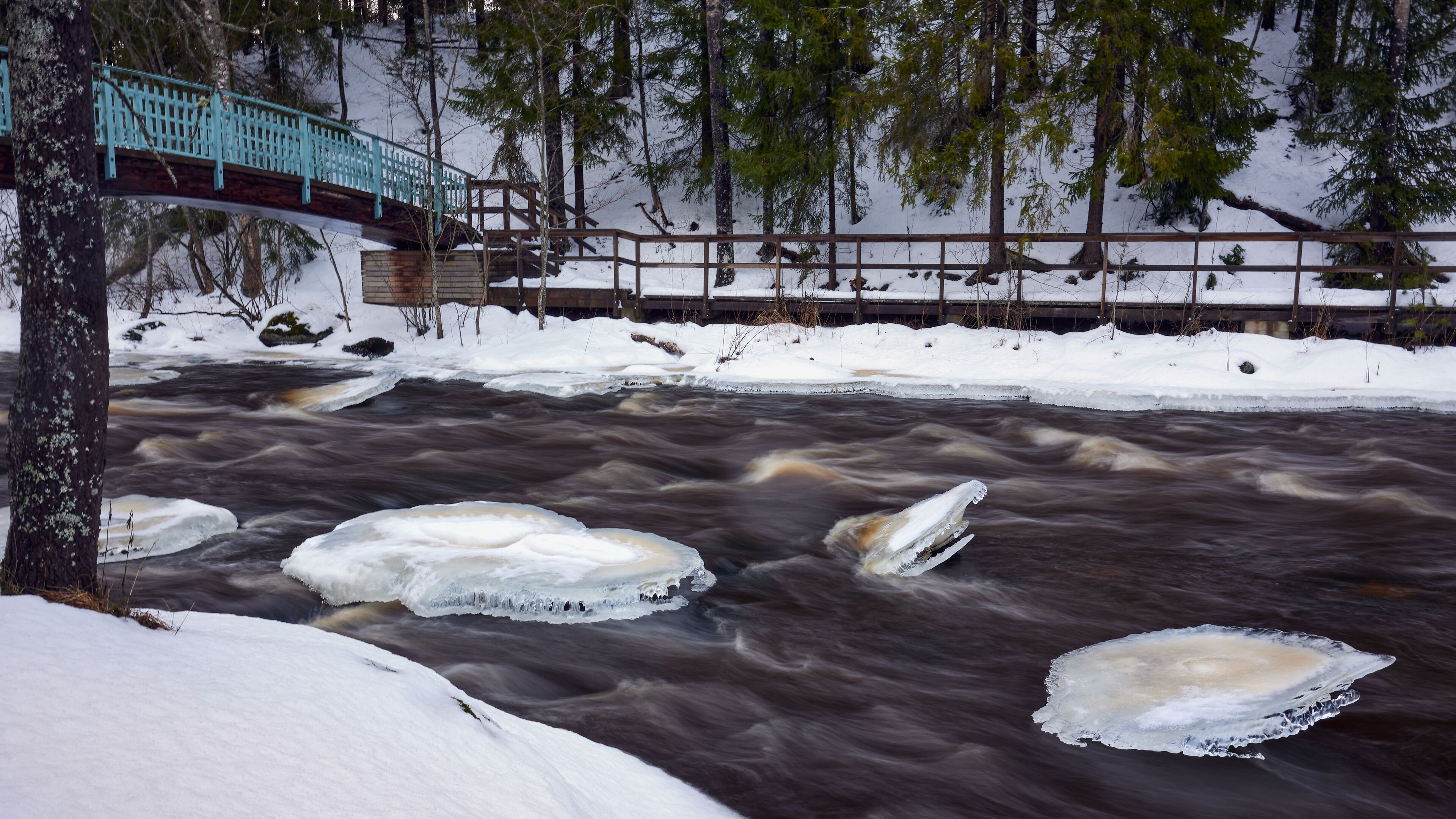 A river with ice floes, a wooden bridge, and snow-covered banks.