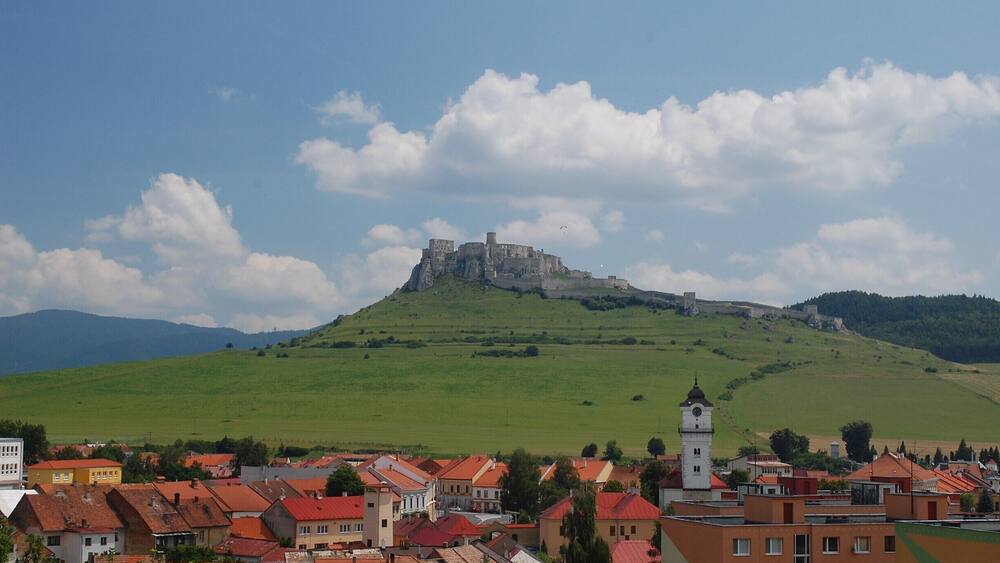 Une ville aux bâtiments aux toits rouges, une colline imposante surmontée d’un château et un ciel d’un bleu limpide.