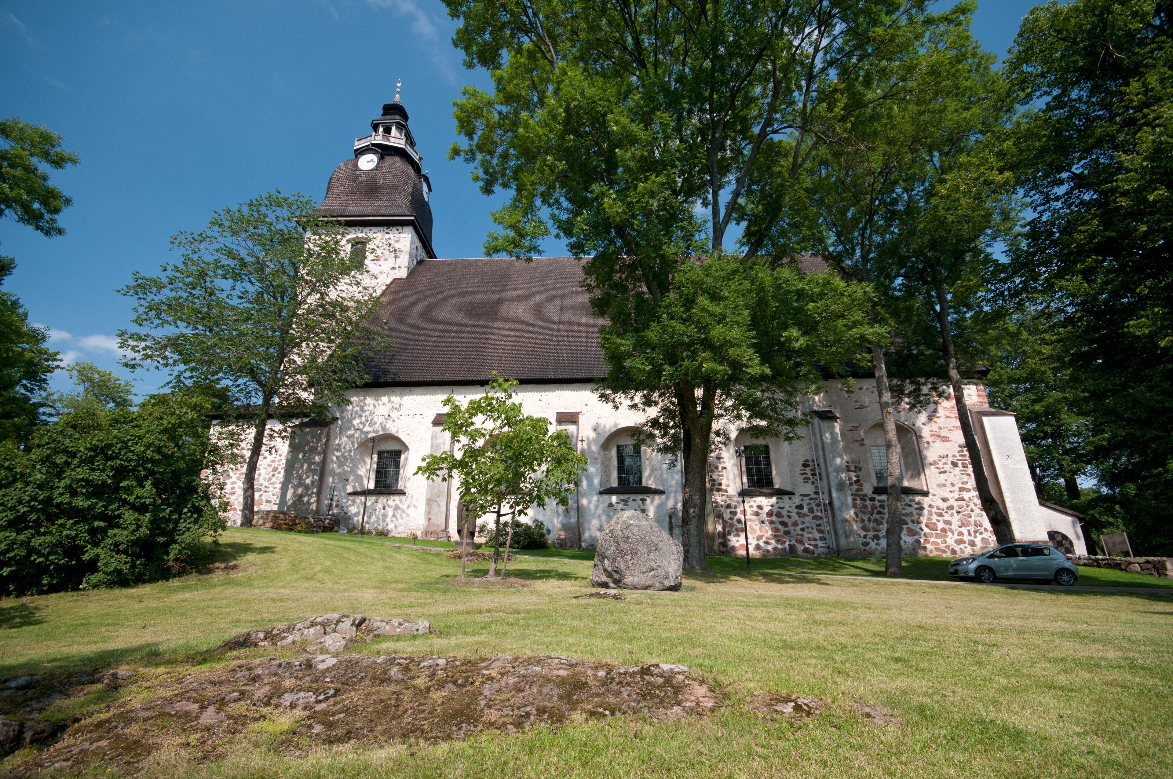 A historic church with a clock tower, surrounded by lush greenery and a stone wall.