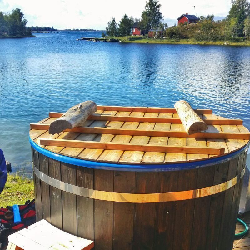 A wooden hot tub with two logs on top, surrounded by a lake and houses in the background.
