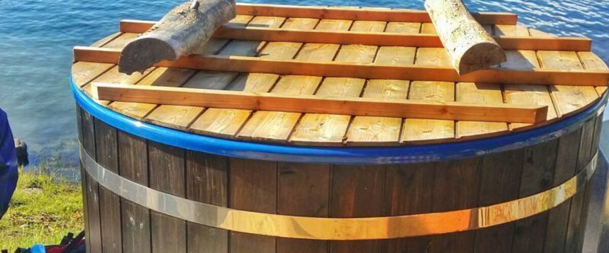 A wooden hot tub with two logs on top, surrounded by a lake and houses in the background.