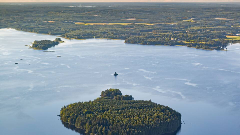 an areial photograph over a lake in finnish countryside near Pielavesi, Finland. The lake is reflecting the light blue of the sky. an island in the foreground, sun is setting