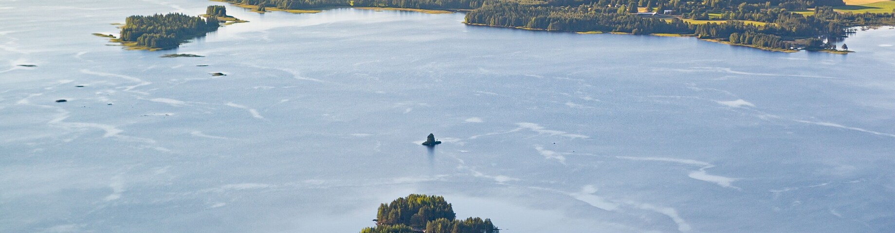 an areial photograph over a lake in finnish countryside near Pielavesi, Finland. The lake is reflecting the light blue of the sky. an island in the foreground, sun is setting