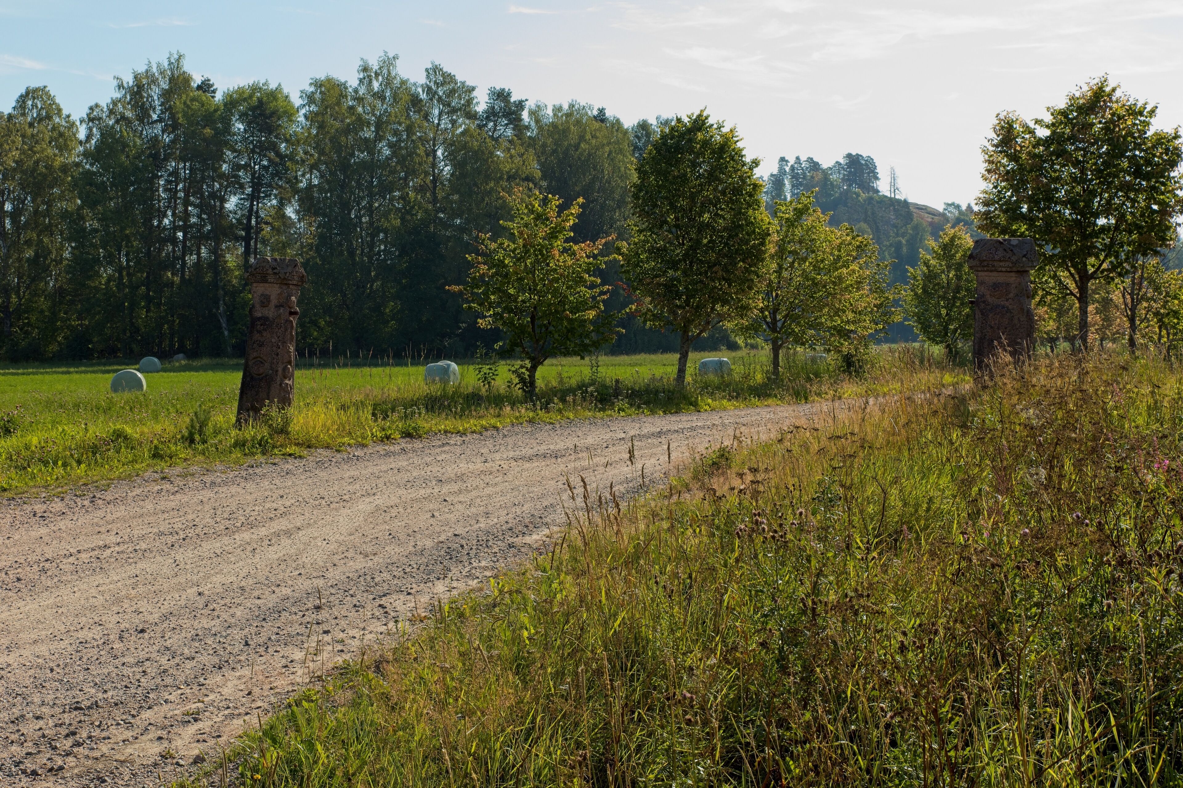 A rural dirt road with hay bales, trees, and stone structures in a field.
