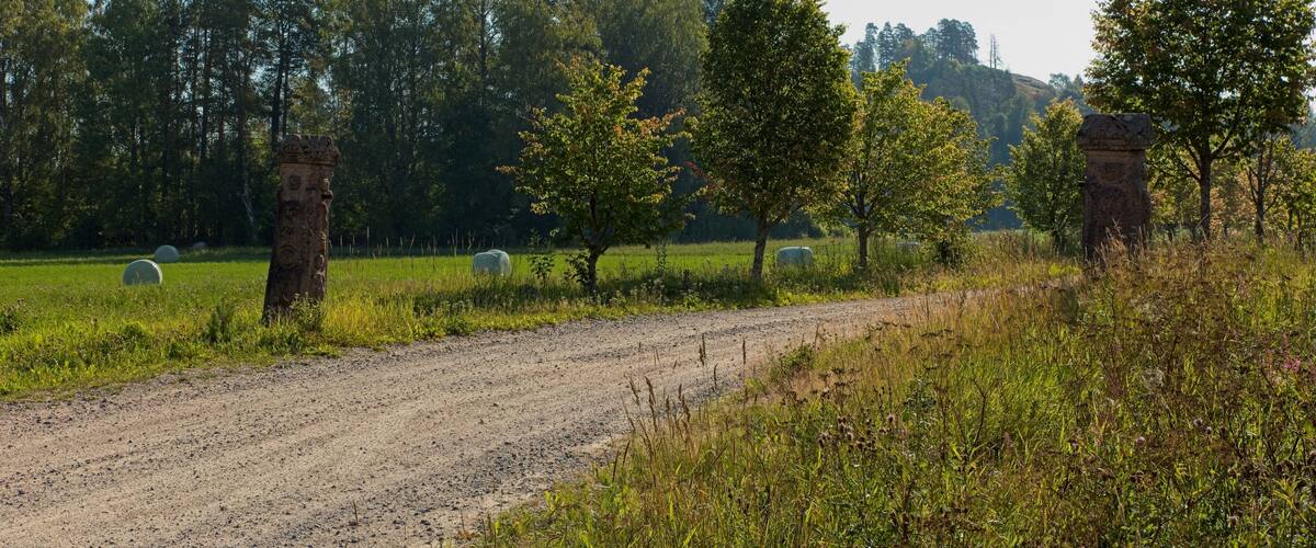 A rural dirt road with hay bales, trees, and stone structures in a field.