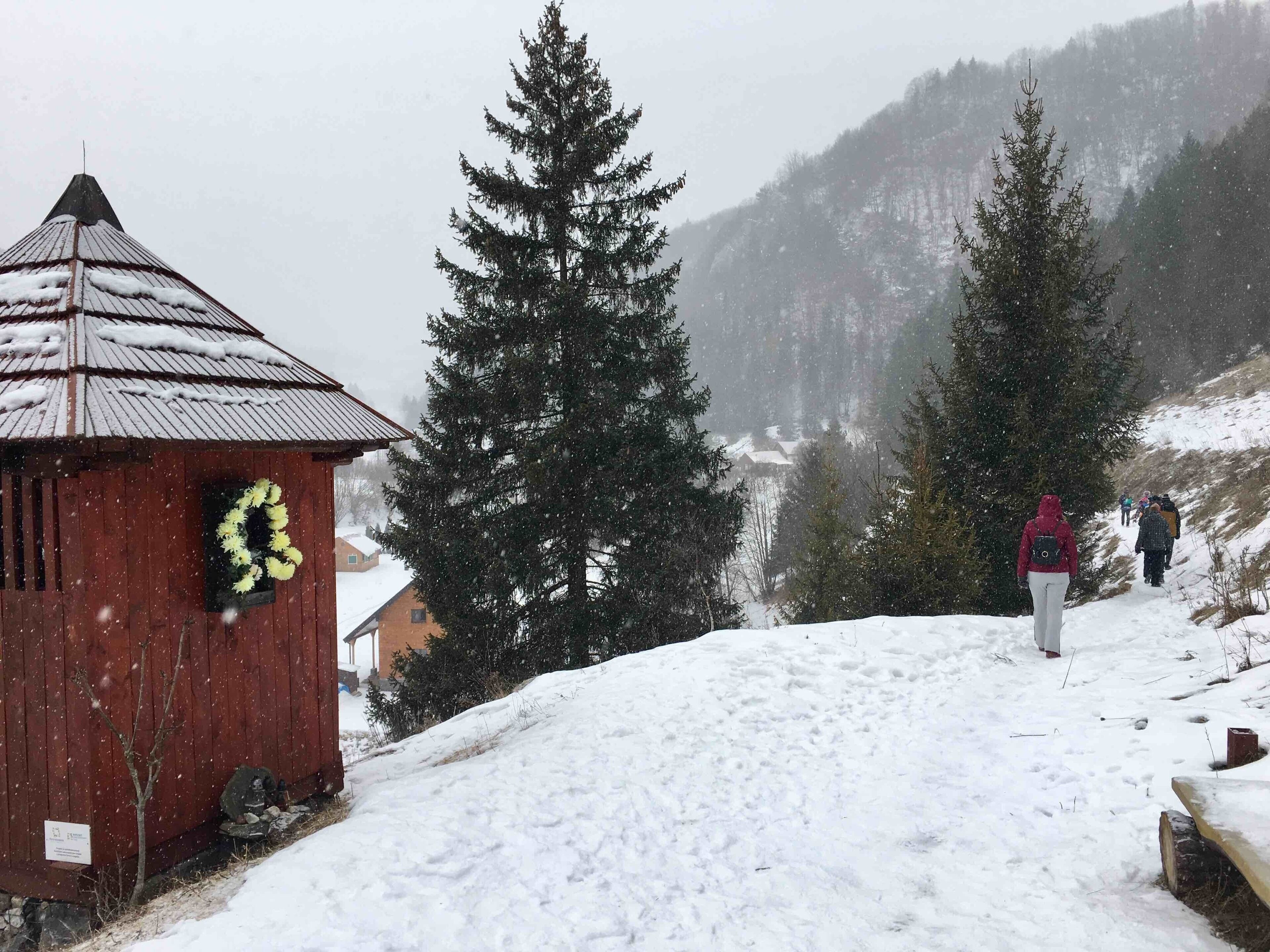 A wooden cabin with a wreath on the door, snow-covered trees, and a snowy mountain landscape.