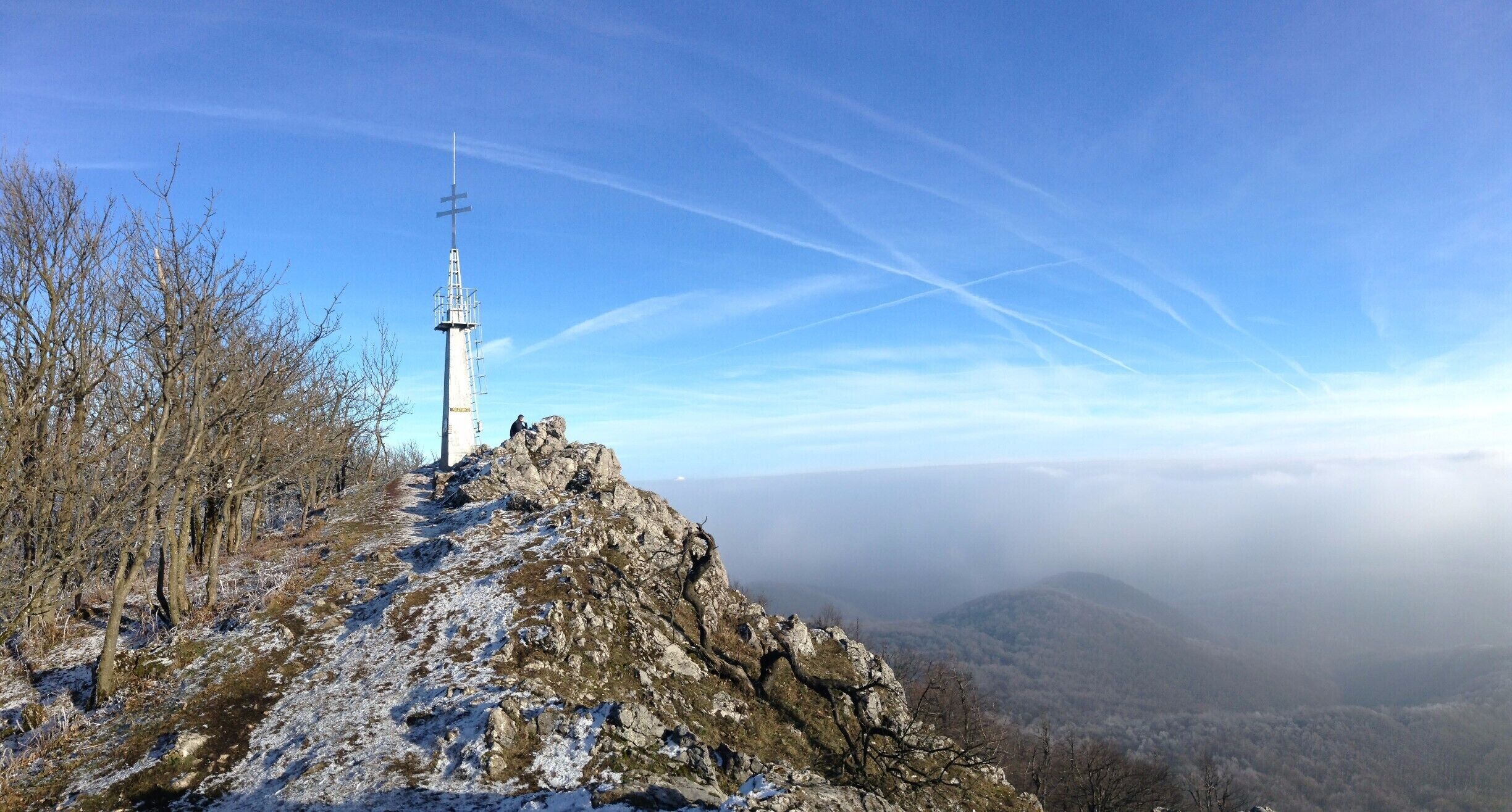 A cross on a rocky hilltop with a view of a valley and distant mountains.