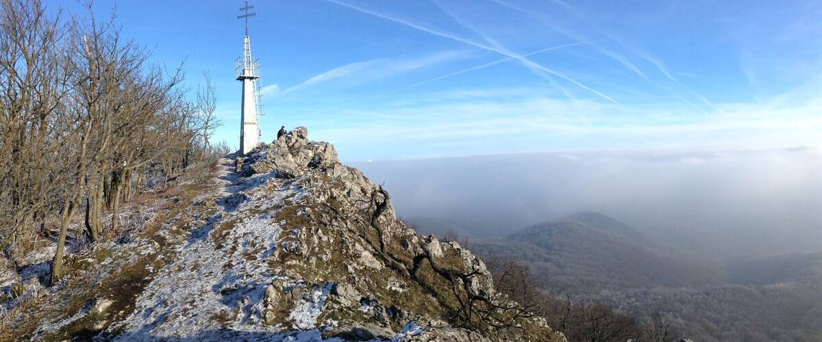 A cross on a rocky hilltop with a view of a valley and distant mountains.