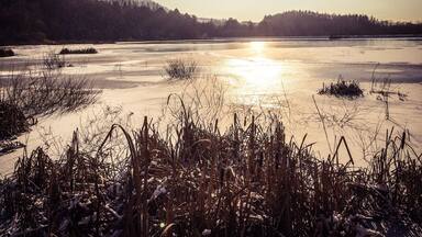 A frozen lake with reflections of the sun, surrounded by hills and sparse vegetation.