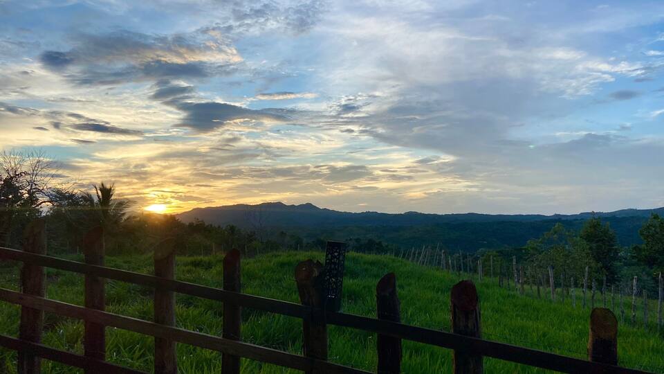 A sunset over a grassy field with a wooden fence in the foreground and mountains in the distance.