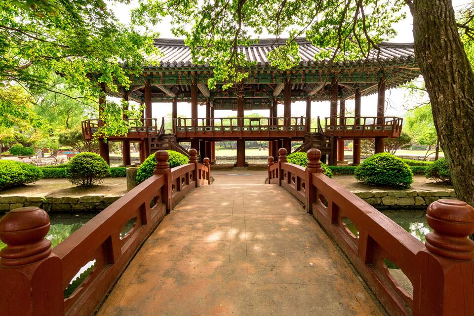 A traditional wooden bridge leading to a pavilion with a tiled roof, surrounded by lush greenery and a stone pathway.