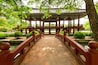 A traditional wooden bridge leading to a pavilion with a tiled roof, surrounded by lush greenery and a stone pathway.
