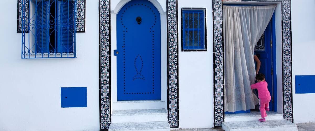 A white building with blue doors and windows, a tiled entrance, and a child in pink.