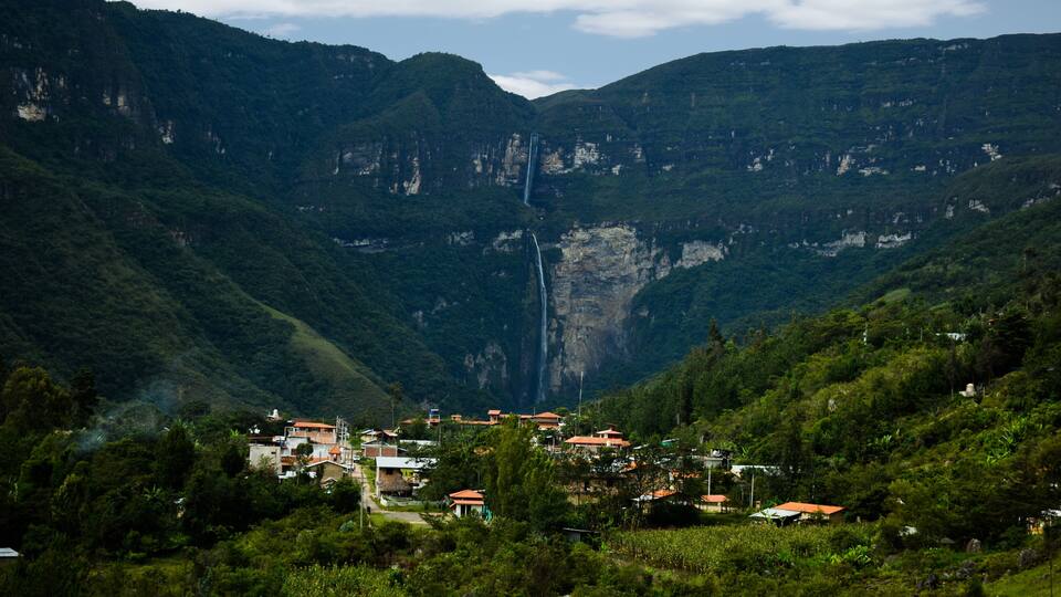 A mountainous landscape with a waterfall, green hills, and a small village with red-roofed houses.