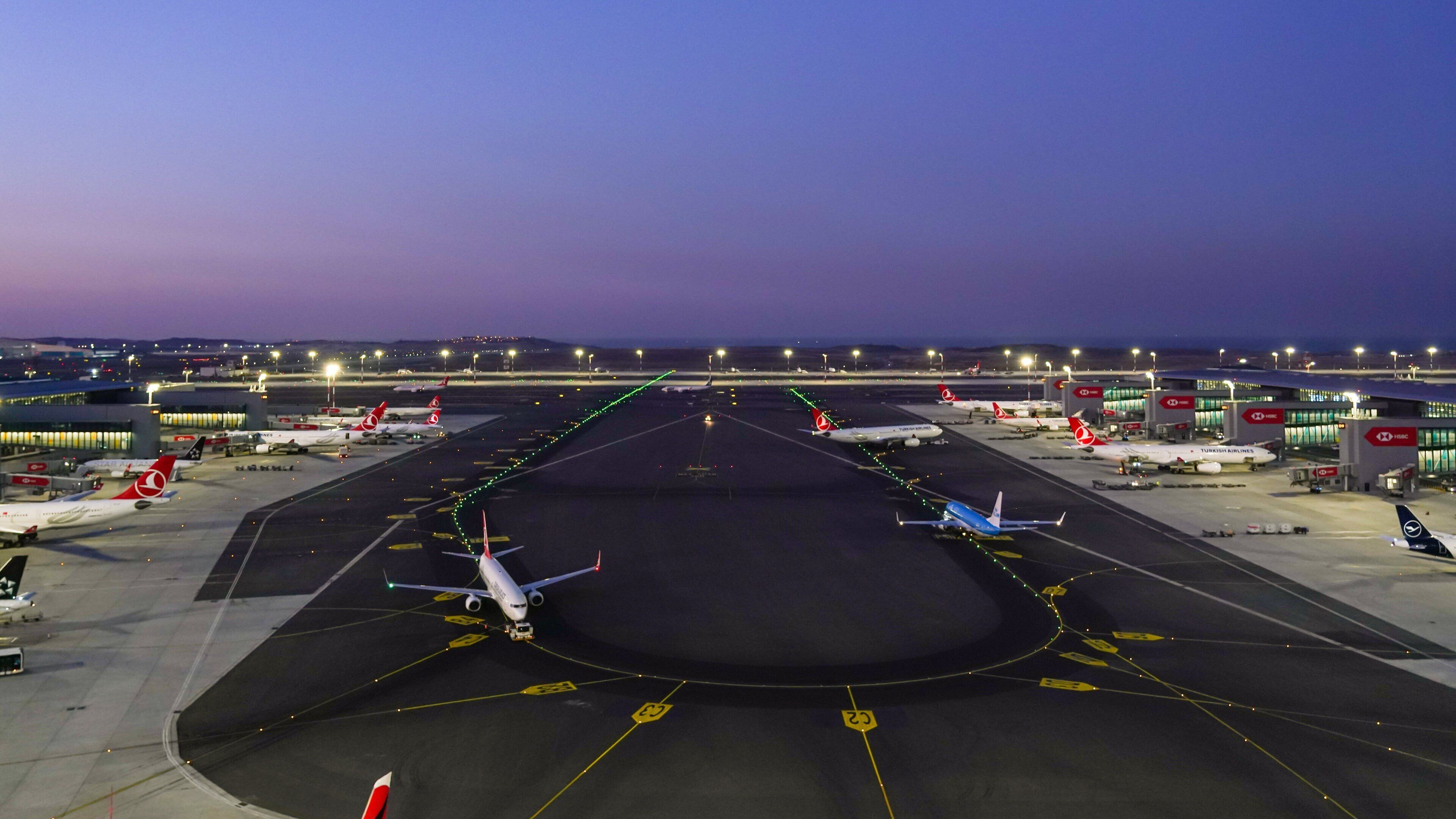 An airport runway with multiple airplanes parked, airport buildings, and a clear sky at dusk.