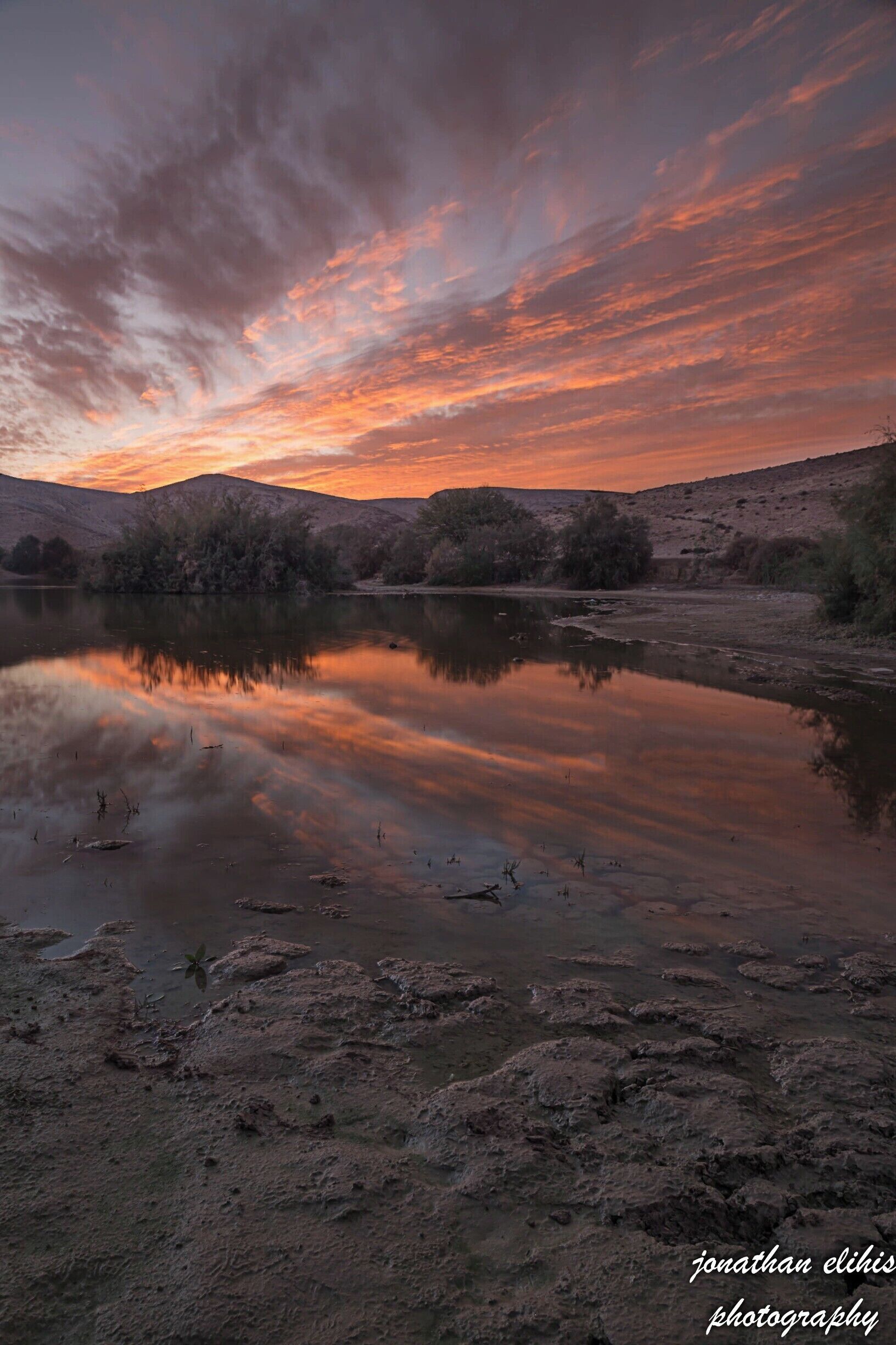 A sunset over a calm lake with reflections of the sky, surrounded by hills and sparse vegetation.