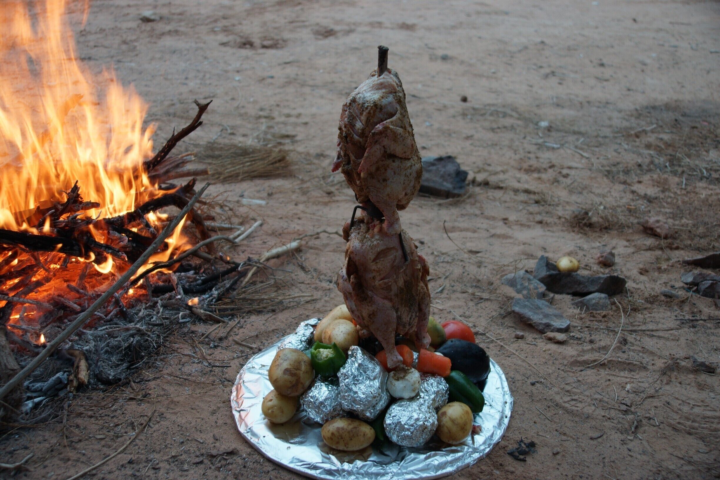 A roasted chicken on a spit over an open fire, surrounded by vegetables on a foil-covered plate.