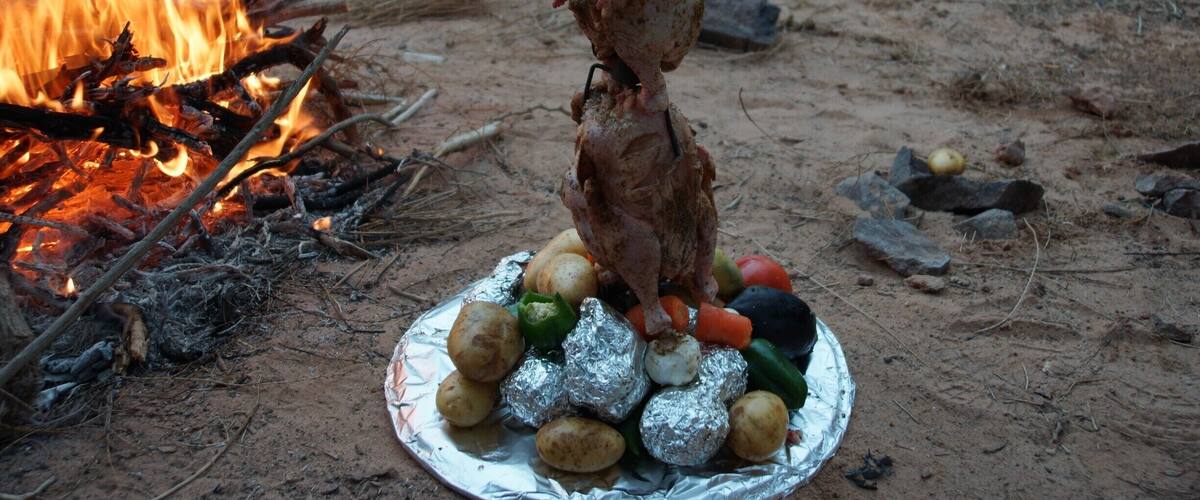 A roasted chicken on a spit over an open fire, surrounded by vegetables on a foil-covered plate.