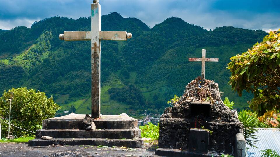 A cross on a stone pedestal with a smaller cross on top, surrounded by lush green hills.