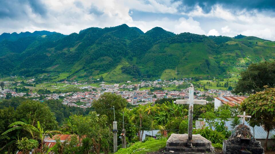 A lush green valley with mountains in the background, a cross monument, and a town below.