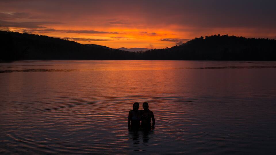 A sunset over a calm lake with silhouettes of two people standing in the water.