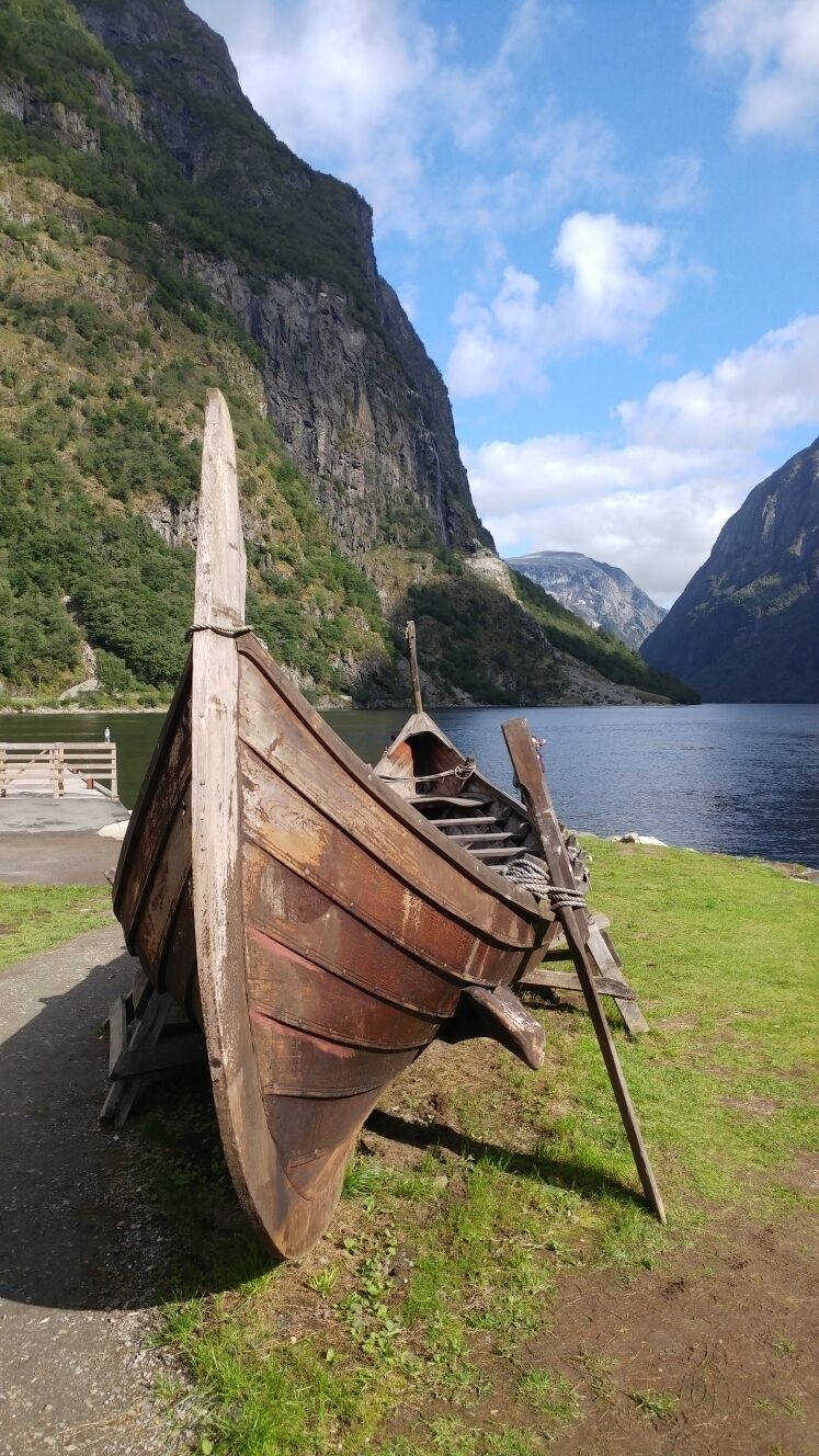 A wooden boat on a grassy shore with a mountainous backdrop and a body of water.