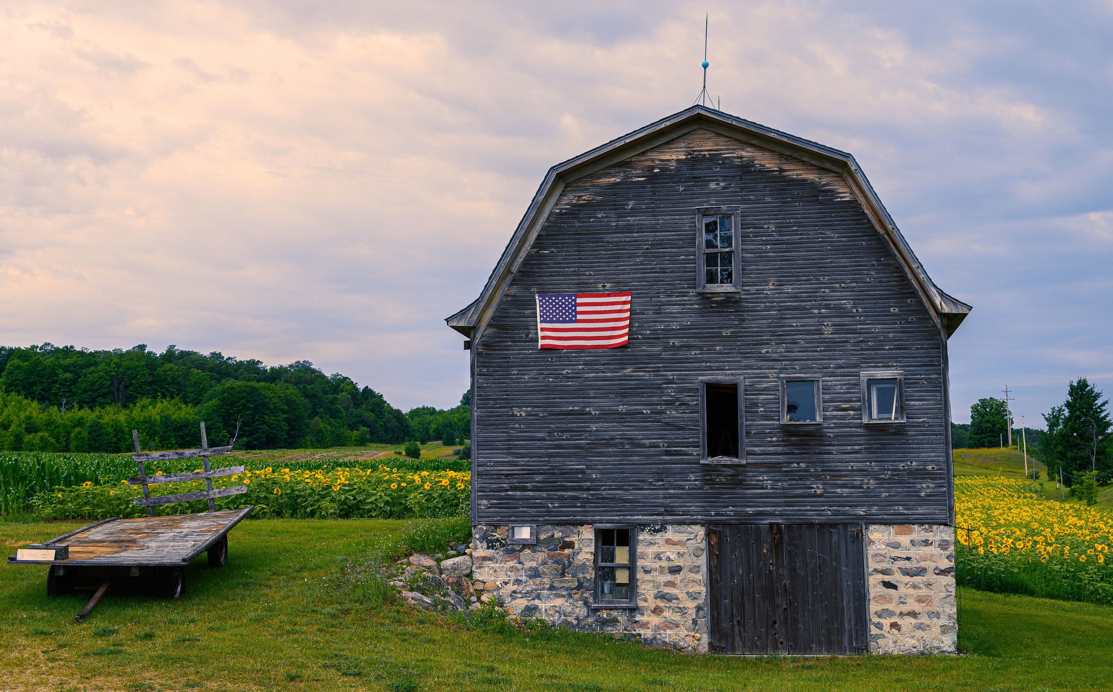 A rustic barn with an American flag, surrounded by a lush green field and a wooden bench.