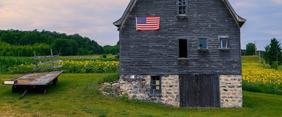 A rustic barn with an American flag, surrounded by a lush green field and a wooden bench.