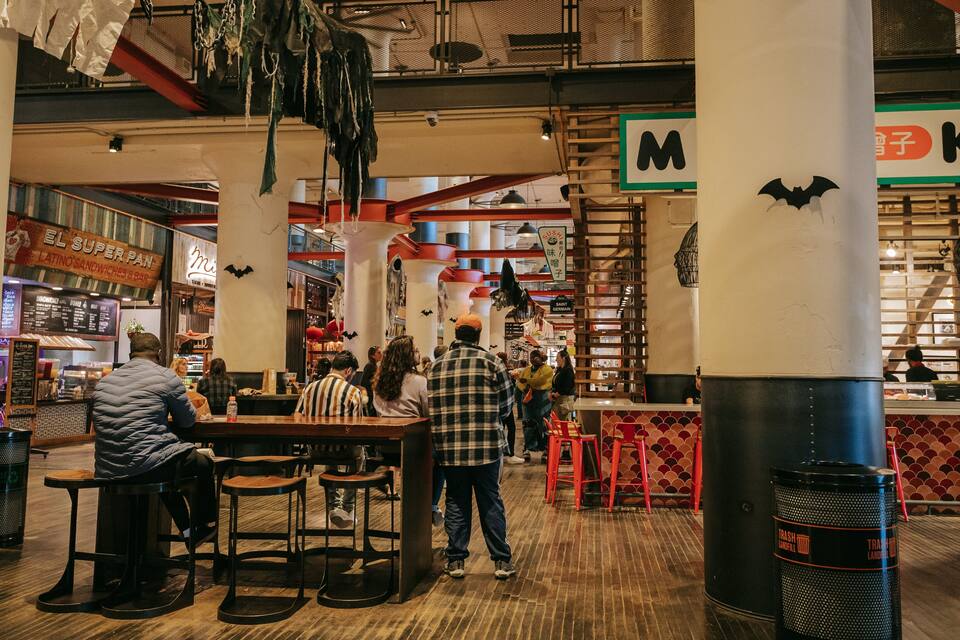 A bustling indoor market with wooden floors, hanging decorations, and various food stalls.
