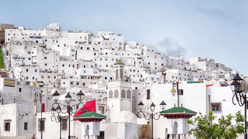 A white-washed hillside town with a prominent red flag, traditional lamp posts, and a mosque with a green dome.
